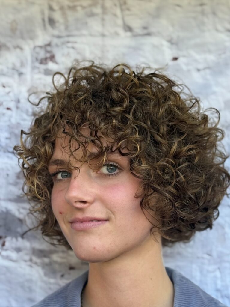 A woman with a chin-length golden brown curly shag and full curly bangs.