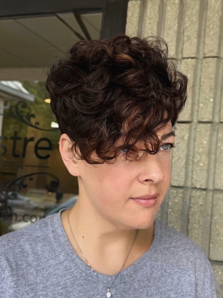 A woman with a short tapered brunette pixie cut featuring voluminous curls and a dramatic side-swept fringe.