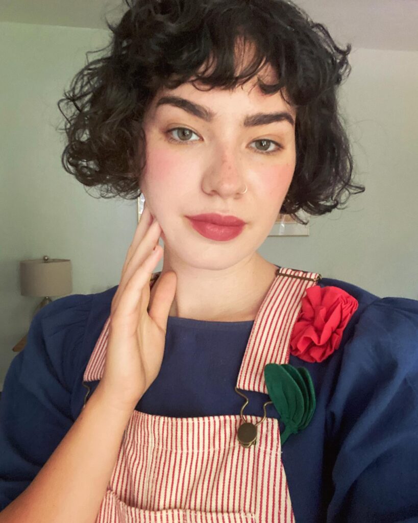 A woman with a dark curly French bob and short microbangs wearing red-striped overalls.