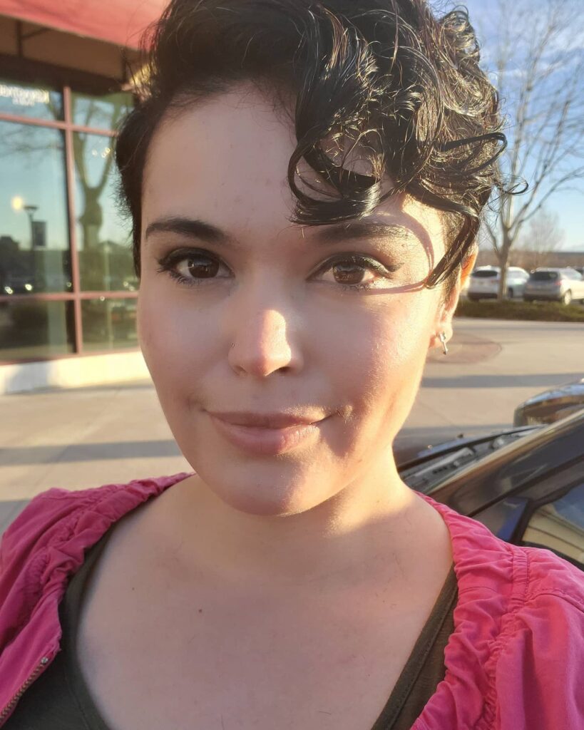 A woman outdoors with a dark asymmetrical pixie cut featuring short sides and long curly bangs resting on her forehead.