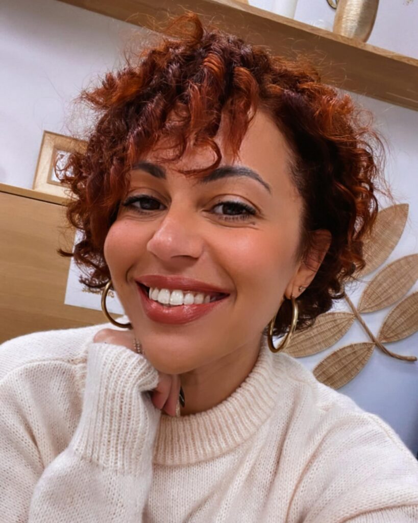 A smiling woman with a short auburn curly pixie cut and bouncy spiral bangs.