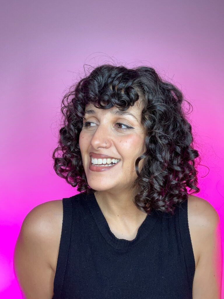 A smiling woman with a mid-length jet black curly bob and thick curly bangs against a pink background.