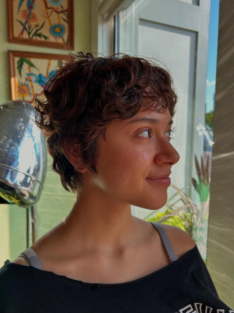 A side profile of a woman with a chestnut brown wavy pixie cut and piecey bangs smiling near a window.