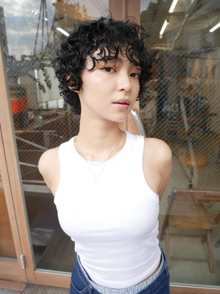 A woman with a short, wet-look, black, curly pixie cut and spiral bangs wearing a white tank top.