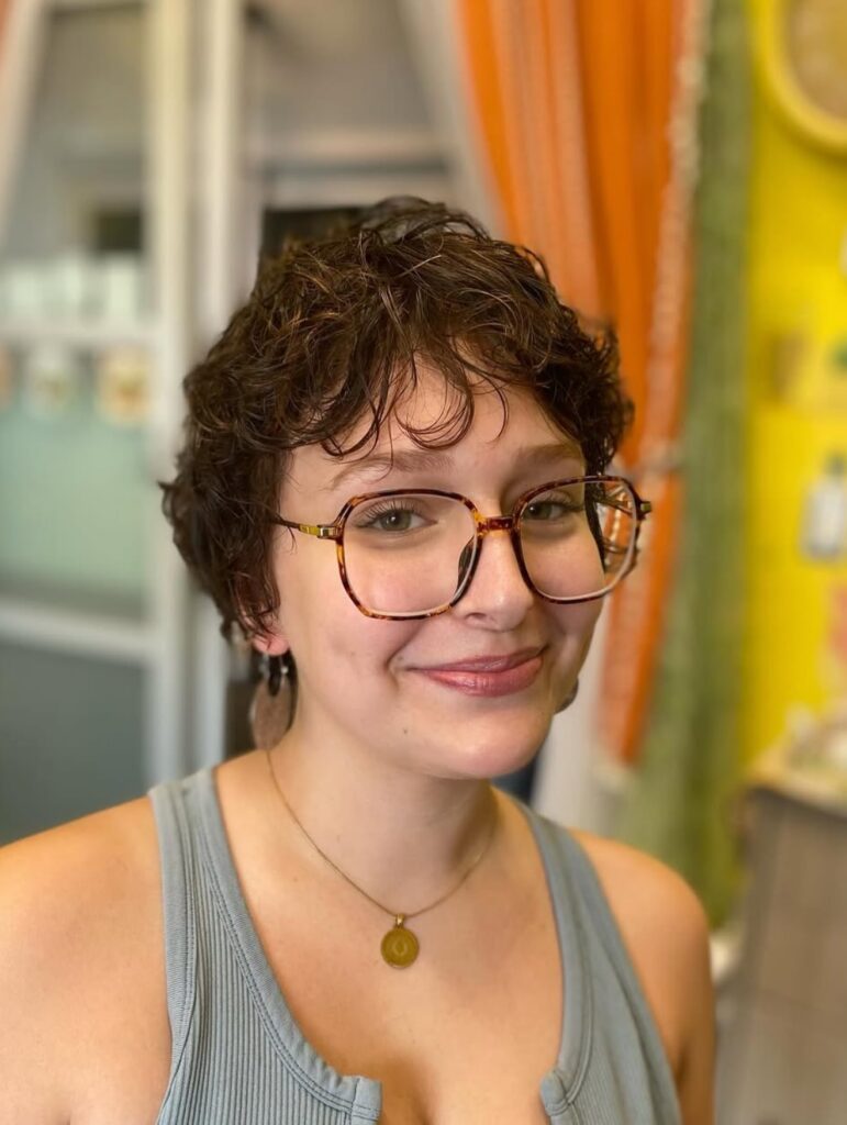 A smiling woman wearing tortoiseshell glasses with a short, wavy brunette pixie cut and wispy bangs.