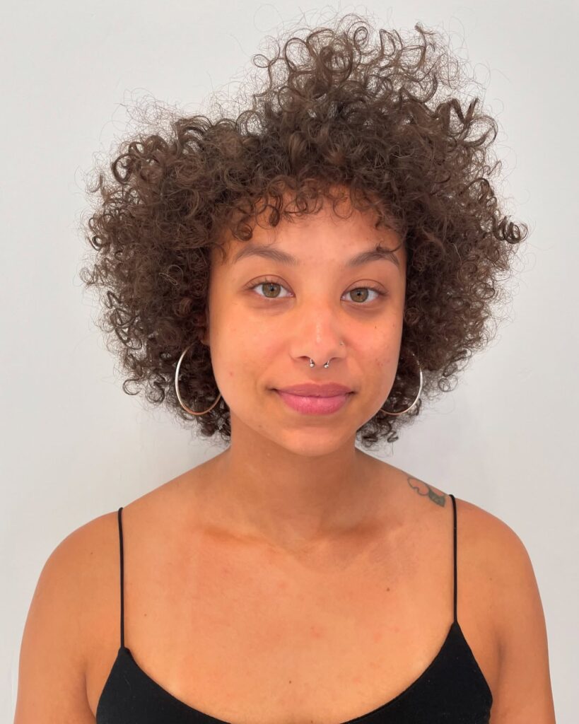 A woman with a voluminous, rounded, short, curly hairstyle in a dark brown color featuring tight coils and soft curly bangs.