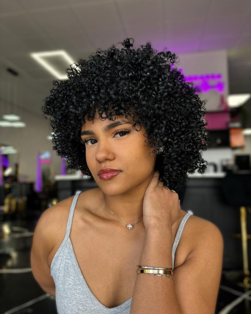 A woman with a voluminous short curly hairstyle in a deep black color with tight defined ringlets and full curly bangs.