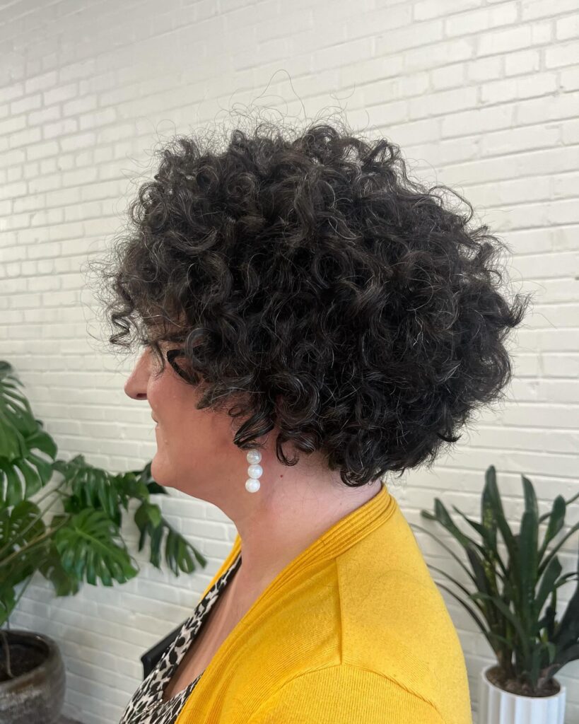 A woman viewed from the side with a short, voluminous, curly wedge haircut in a salt-and-pepper color.