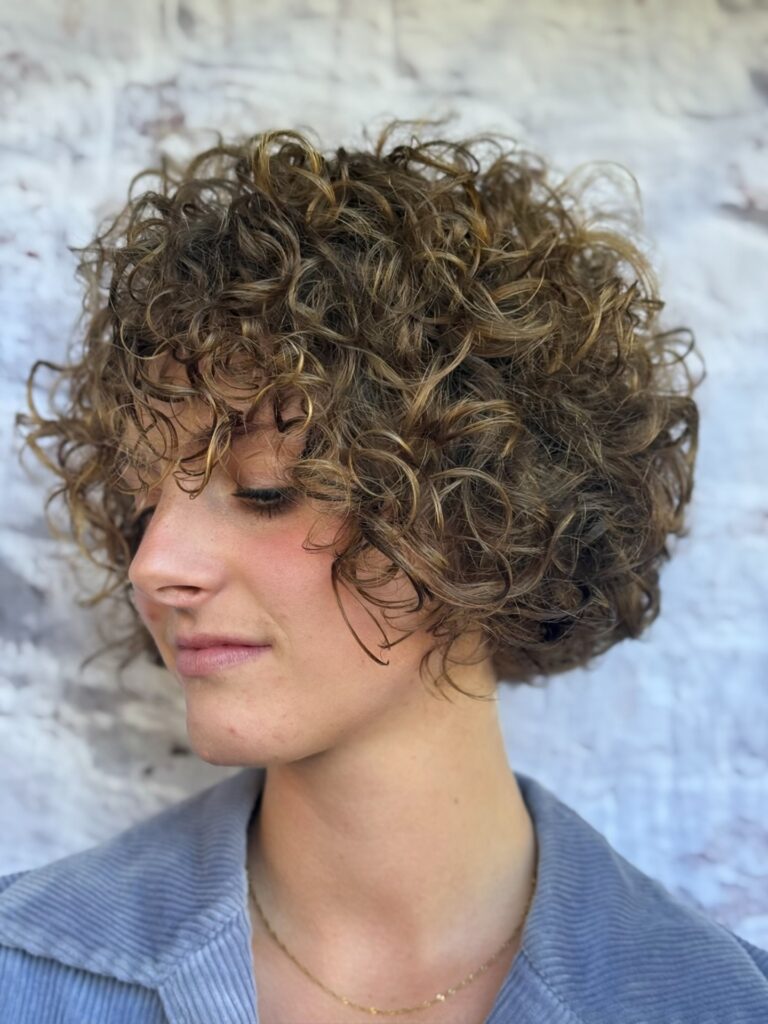 A woman with a short, rounded, curly bob in a light brown color with blonde highlights and soft, wispy, curly bangs.