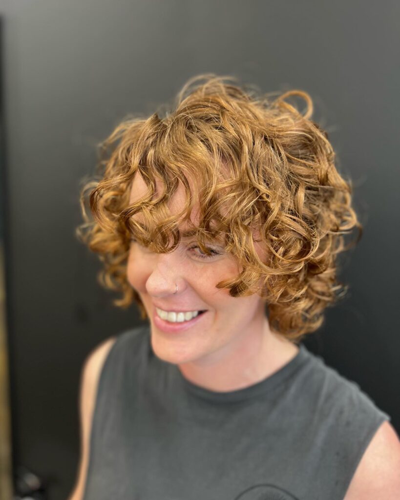 A smiling woman with a voluminous short curly shag hairstyle in a bright copper ginger color.