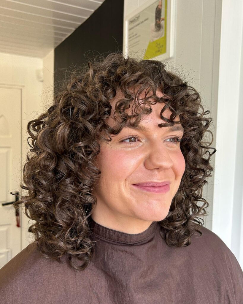 A woman with a voluminous short curly shag hairstyle in a glossy brunette color with thick, defined ringlets and curly bangs.
