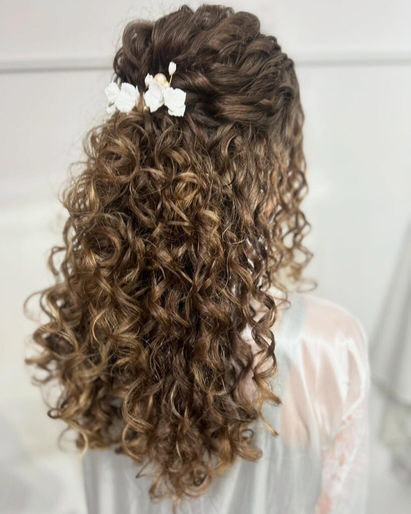 A woman with long curly brown hair styled in a half updo decorated with small white flowers.