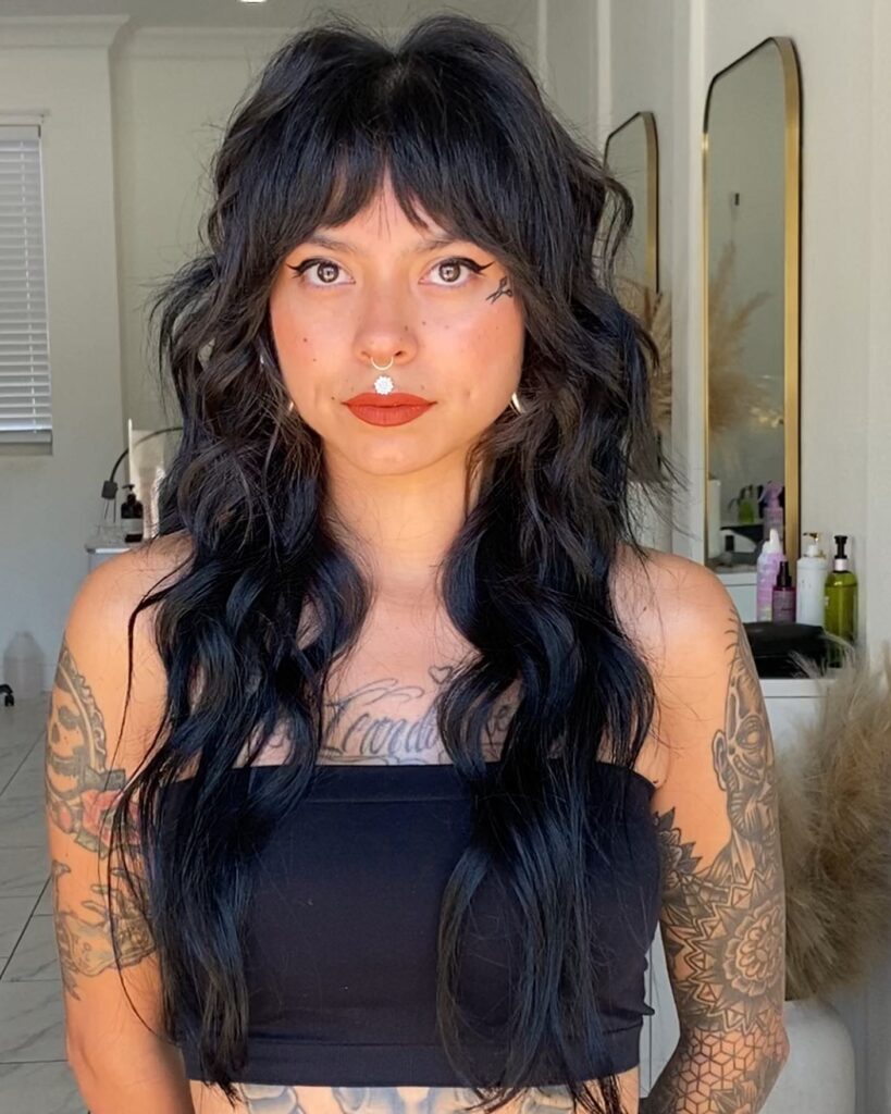 A woman with long wavy jet black hair styled into a heavy shag with textured bangs, facial piercings, and arm tattoos.