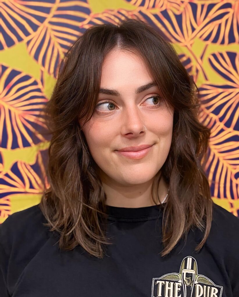 A woman with shoulder length light brown hair styled in a textured shag with center parted curtain bangs and subtle blonde highlights.