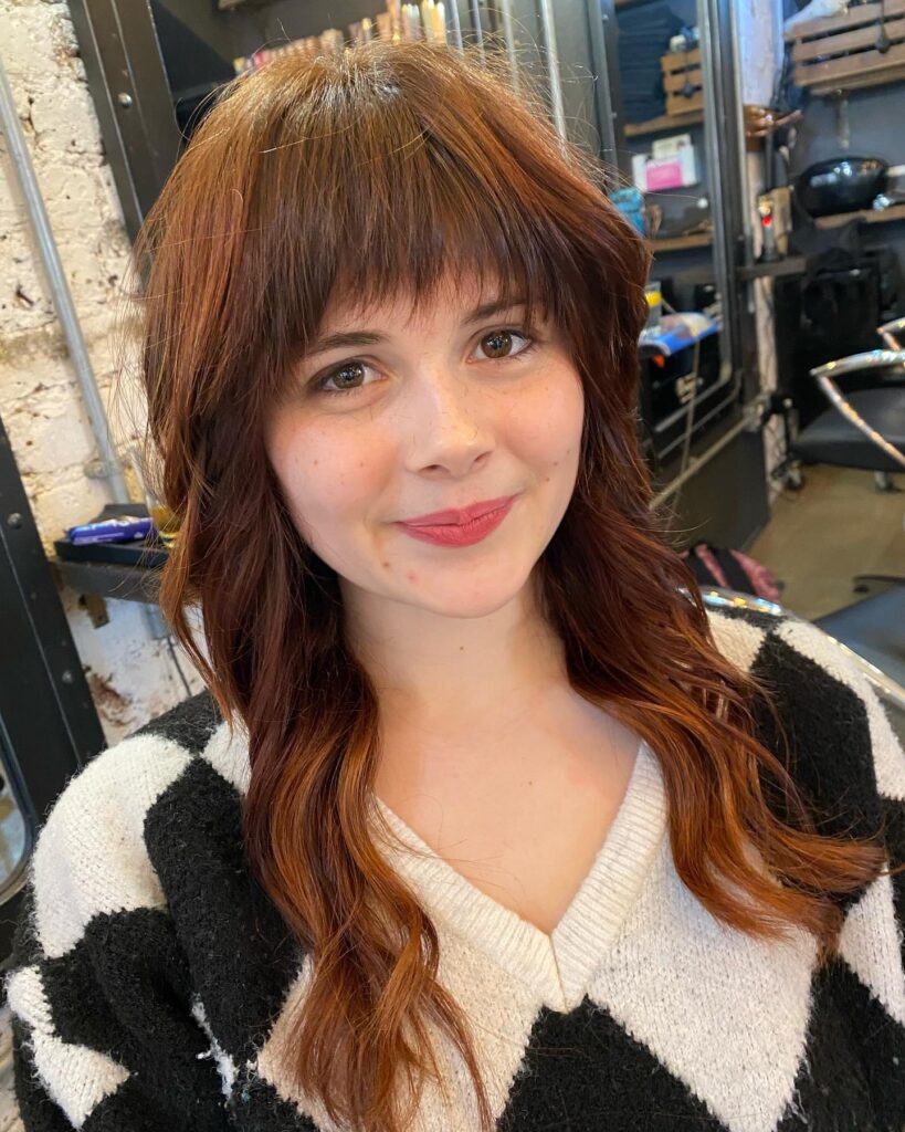 A young woman with medium length auburn hair styled in a layered shag cut with full straight bangs and soft waves.