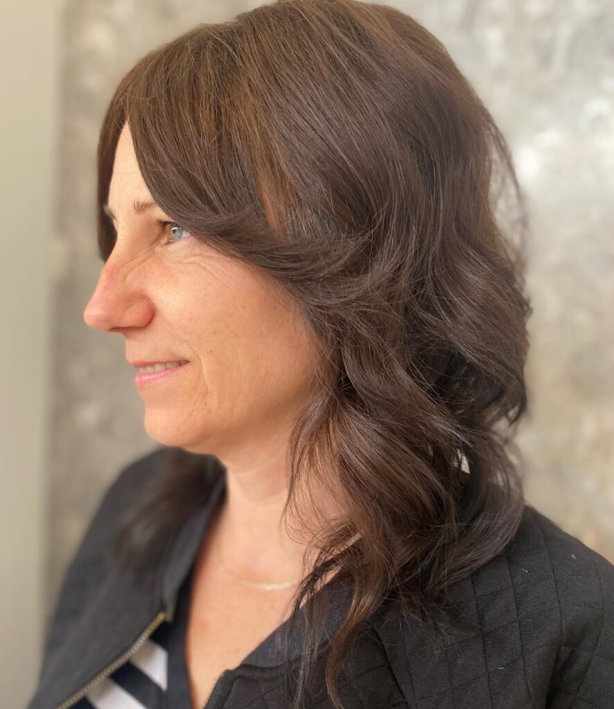 A side profile of a woman with medium length chocolate brown hair styled in a layered wavy shag with long curtain bangs.
