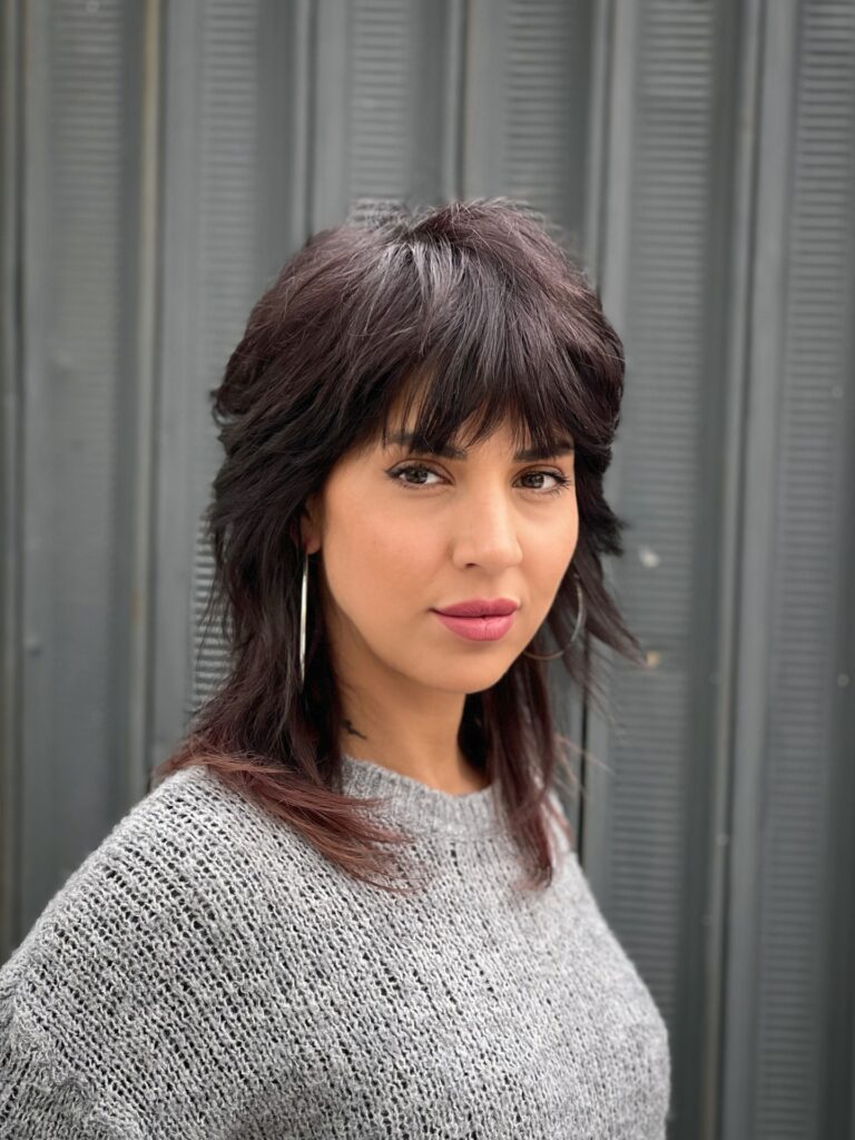 A woman with a dark brunette shag haircut featuring choppy layers and thick textured bangs posing in front of a grey industrial background.