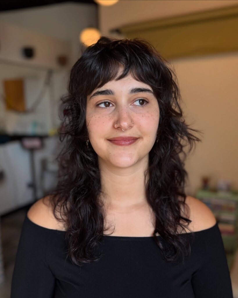 A portrait of a woman with long wavy dark brunette hair in a messy layered shag haircut with baby bangs and facial piercings.
