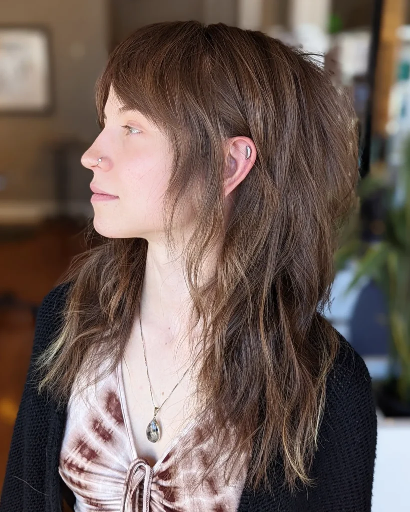 A side profile of a woman with long mousy brown hair in a heavily layered shag haircut with long bangs tucked behind her ear.