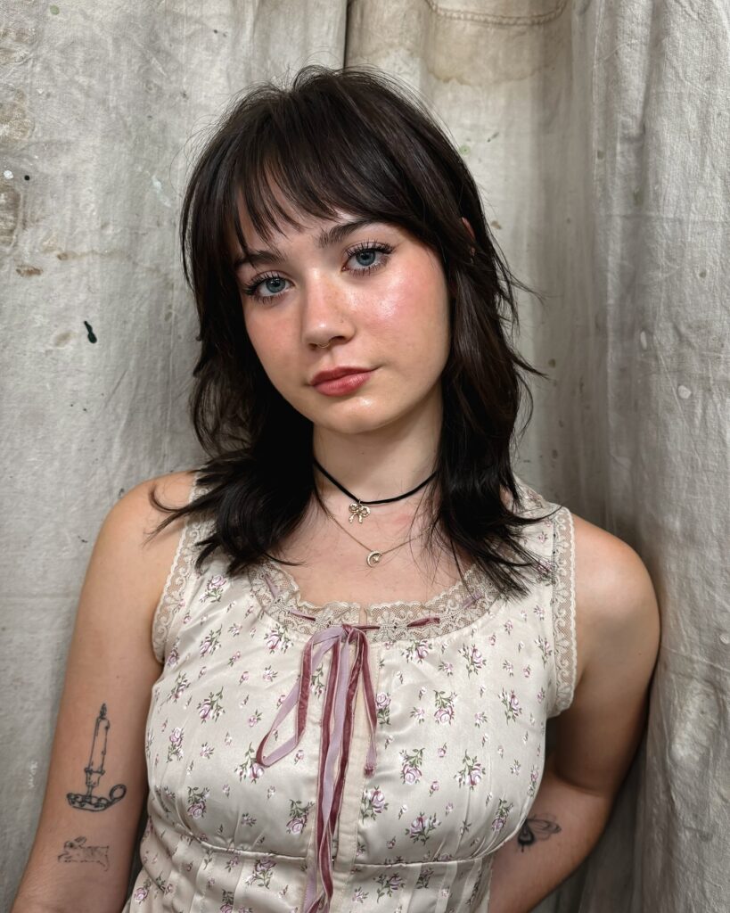 A woman with medium length dark brunette hair in a feathery shaggy haircut with wispy bangs and flicked out layered ends.