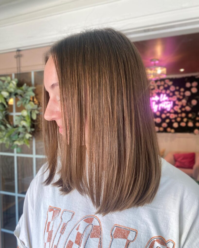 A woman with a straight medium length honey brown haircut featuring a blunt finish and subtle highlights.