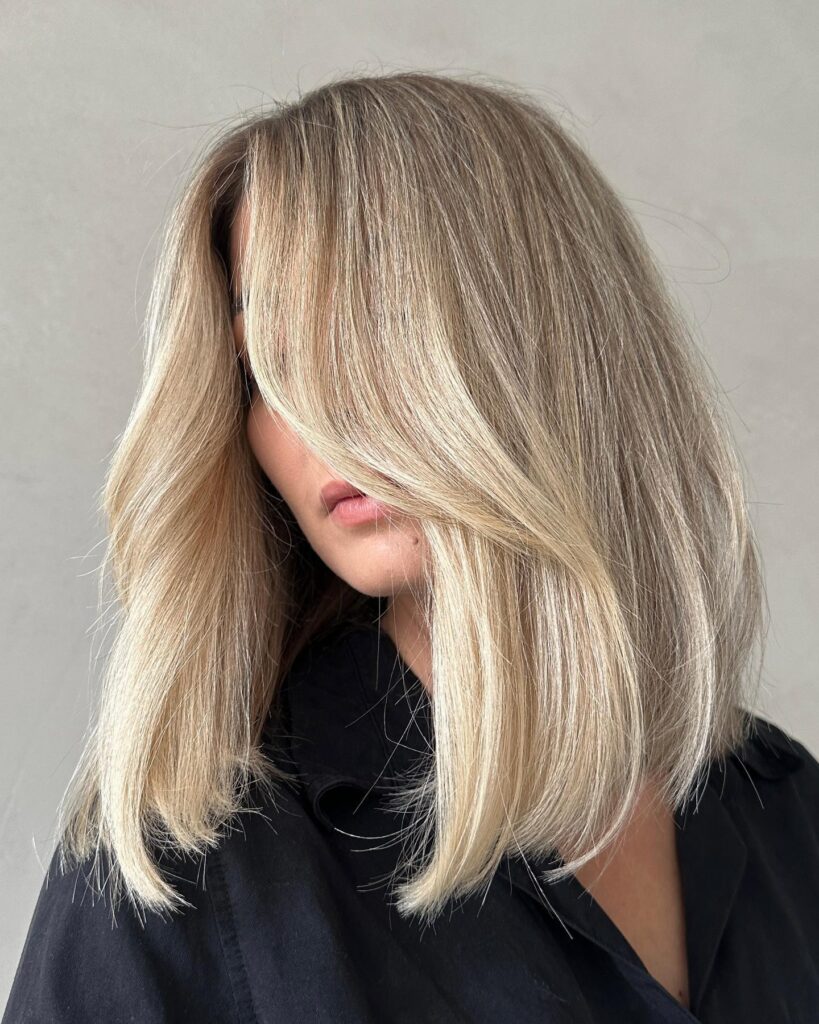 A woman with a medium length sandy blonde lob featuring soft face framing layers and a smooth blowout style.