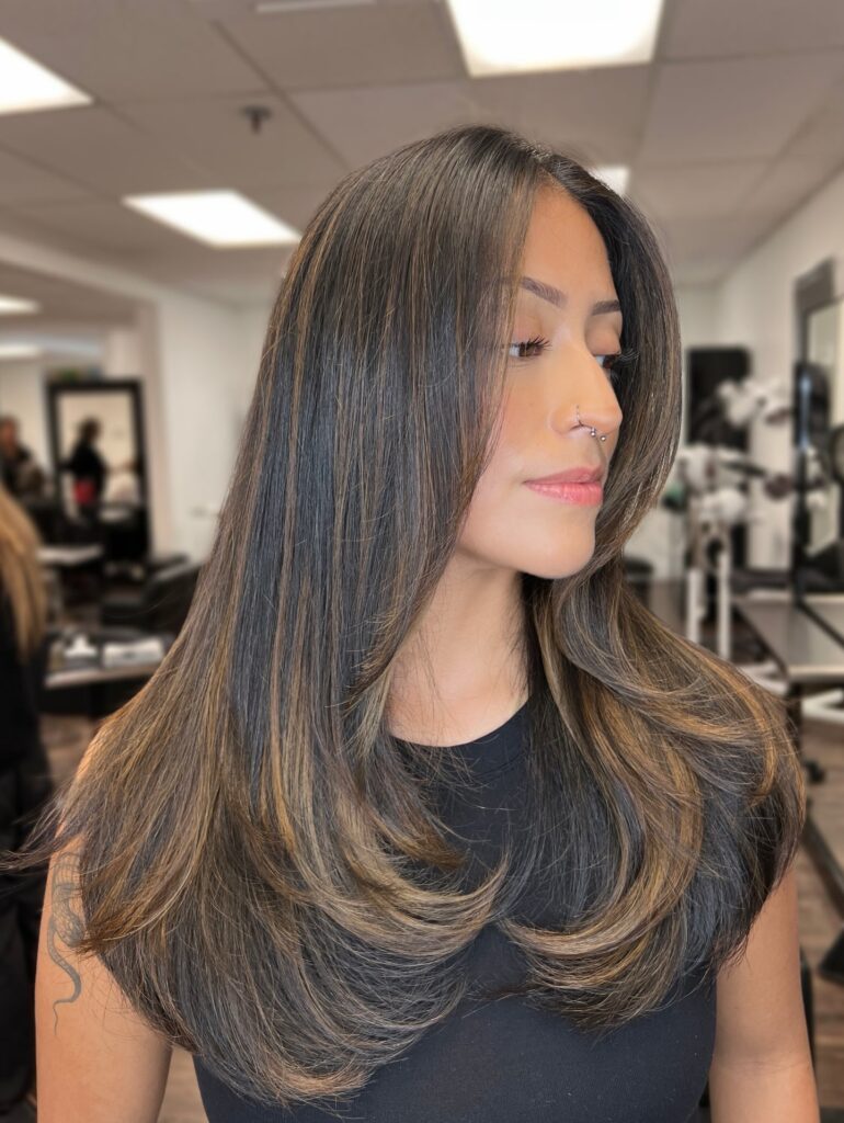 A woman with long brunette hair featuring caramel highlights and voluminous layered ends curved inward.