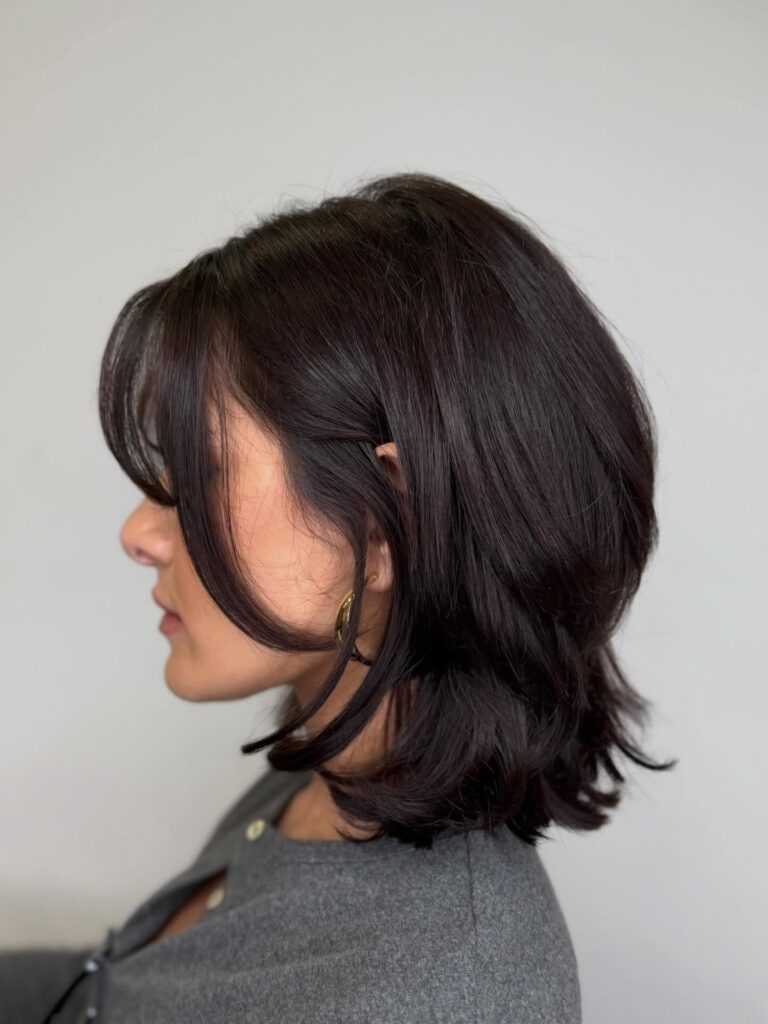 A woman with a dark brunette medium length shaggy haircut featuring wispy bottleneck bangs and textured layers.