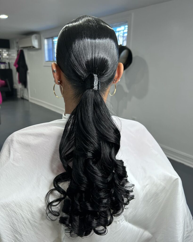 A back view of a woman featuring a slicked back low ponytail that transitions into voluminous dark bouncy curls.