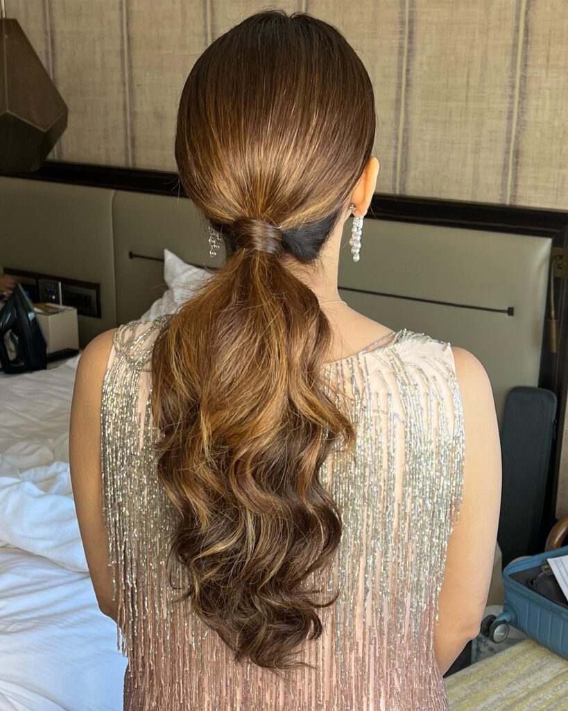 A back view of a woman with long wavy copper hair styled into a polished low ponytail with a hair wrapped base.
