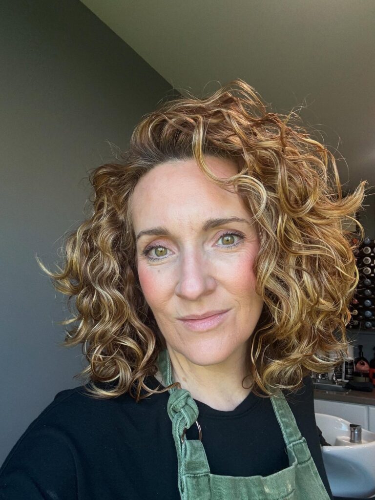 A woman with shoulder-length curly hair featuring copper and golden blonde highlights and voluminous layers.
