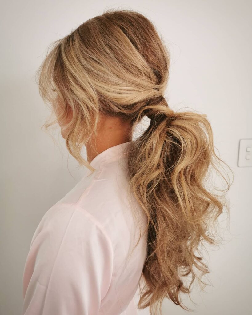 A side profile view of a woman with long, voluminous, wavy blonde hair styled in a low, messy ponytail.