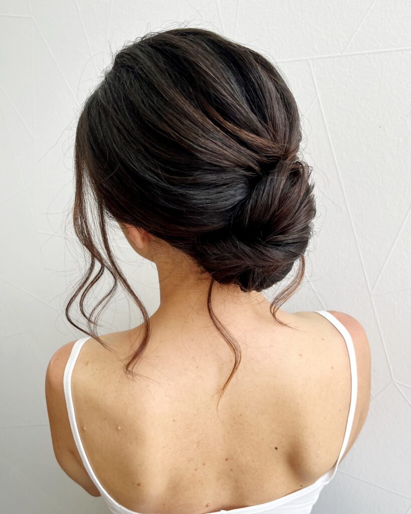 A rear view of a woman with dark brown hair styled in a graceful low chignon with loose curled tendrils framing her neck.