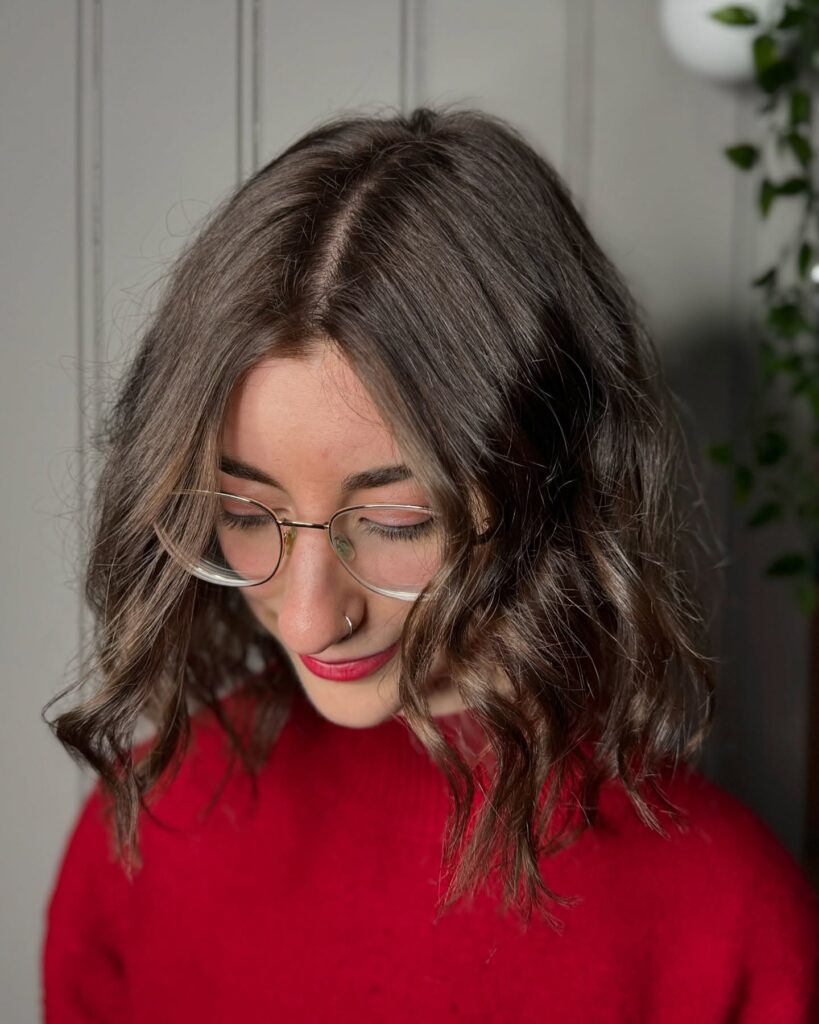 A young woman with shoulder-length dark brown hair with subtle light highlights, styled in loose waves and framed around her face with gold-rimmed glasses.