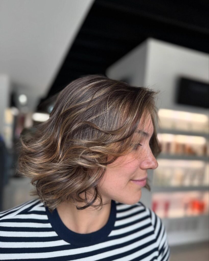 A woman in profile with shoulder-length brunette hair featuring caramel highlights and soft, voluminous waves.