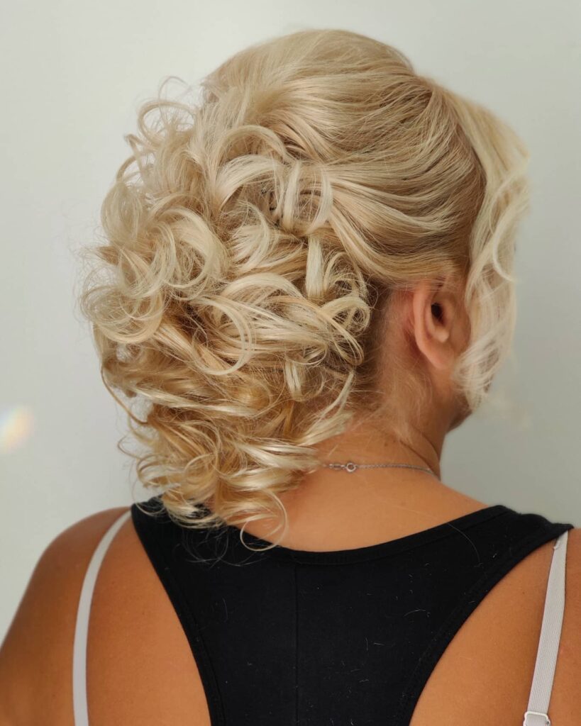 A rear view of a woman with champagne-blonde hair styled in a voluminous curly updo with loose ringlets pinned at the back.