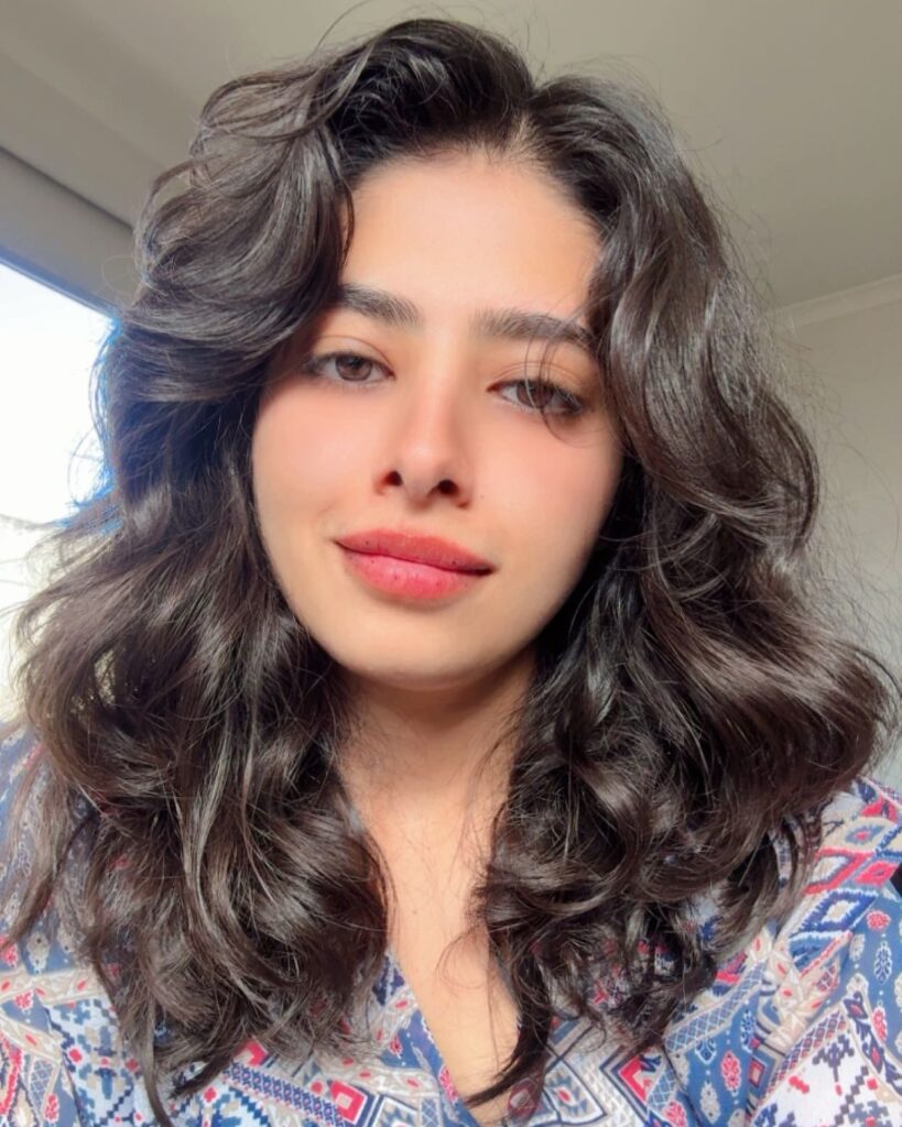 A woman with voluminous shoulder-length dark wavy hair featuring thick face-framing layers and a soft side-swept fringe.