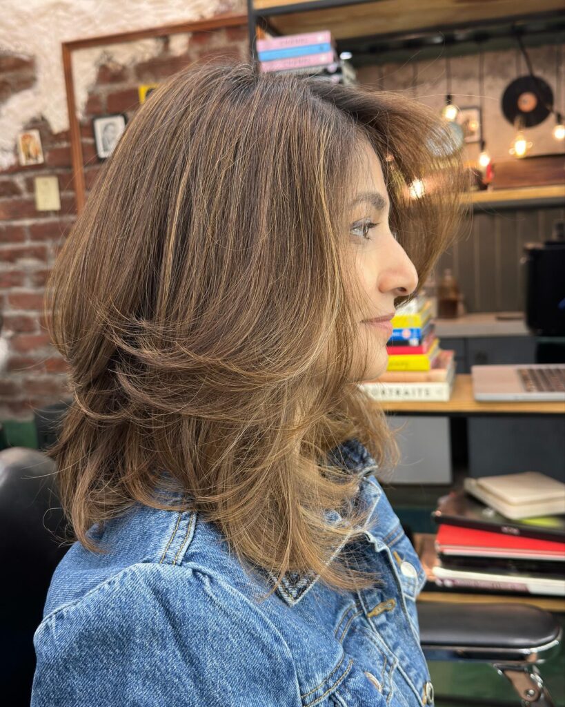 A side profile of a woman with shoulder-length toffee brown hair featuring feathered layers that sweep back away from the face.