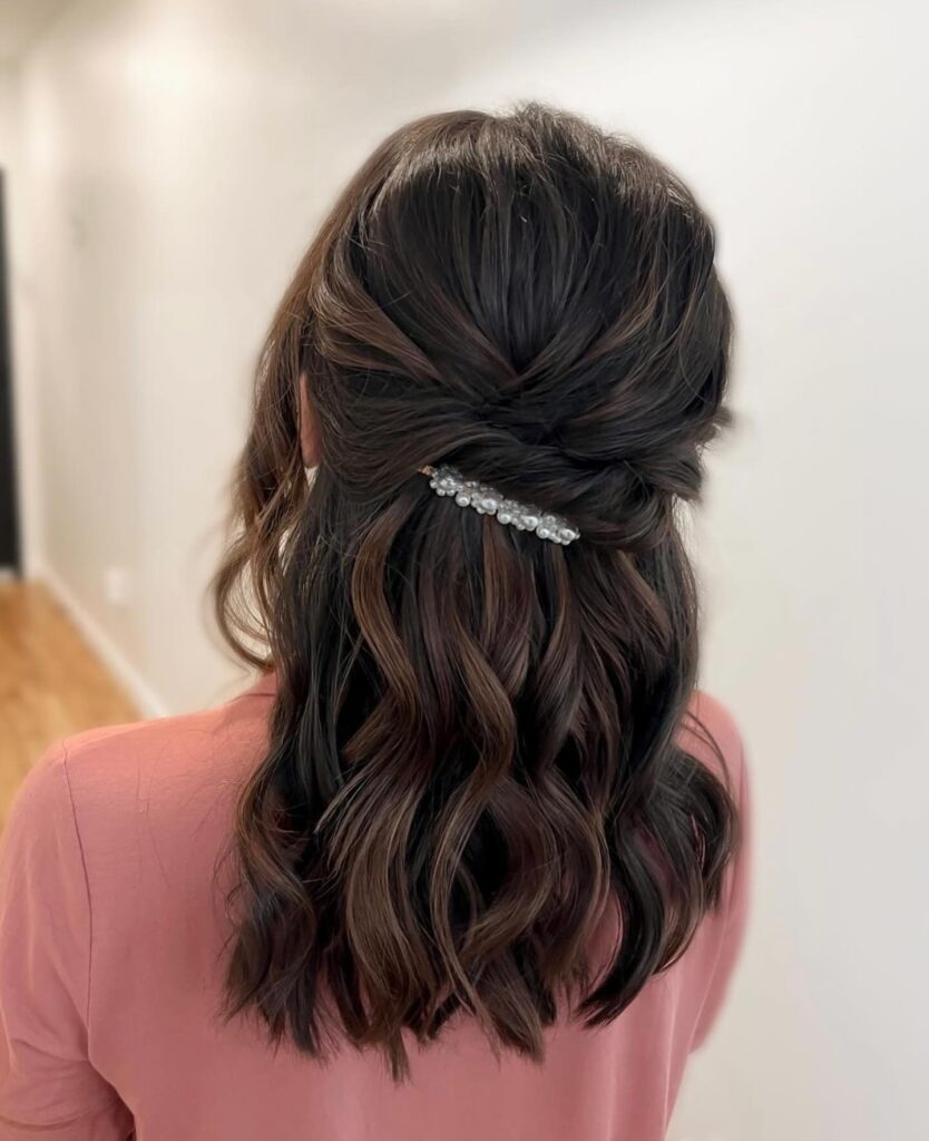 A rear view of a woman with shoulder-length dark brown wavy hair styled in a half-up twisted look and secured with a white pearl barrette.