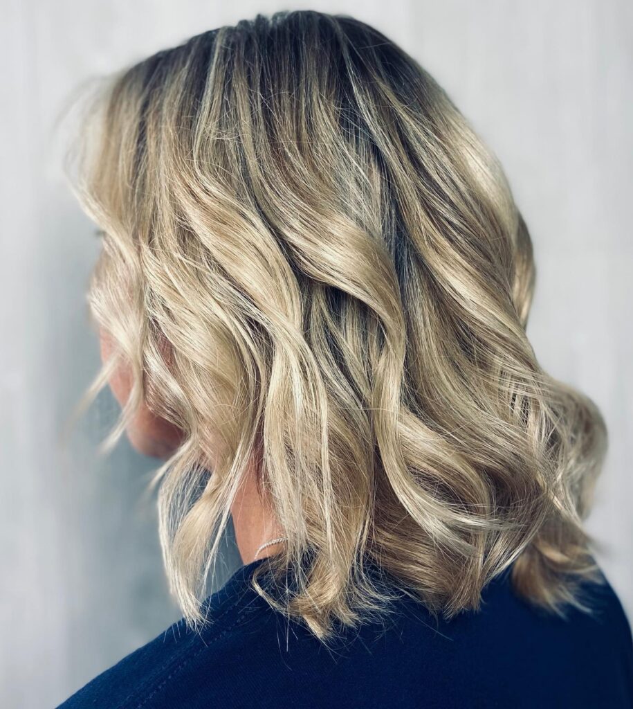 A rear-side view of a woman with shoulder-length ashy blonde hair styled in loose beachy waves with darker roots.
