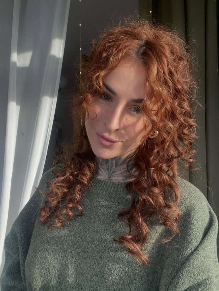 A woman with medium-length, vibrant, curly copper hair and soft ringlets framing her face.