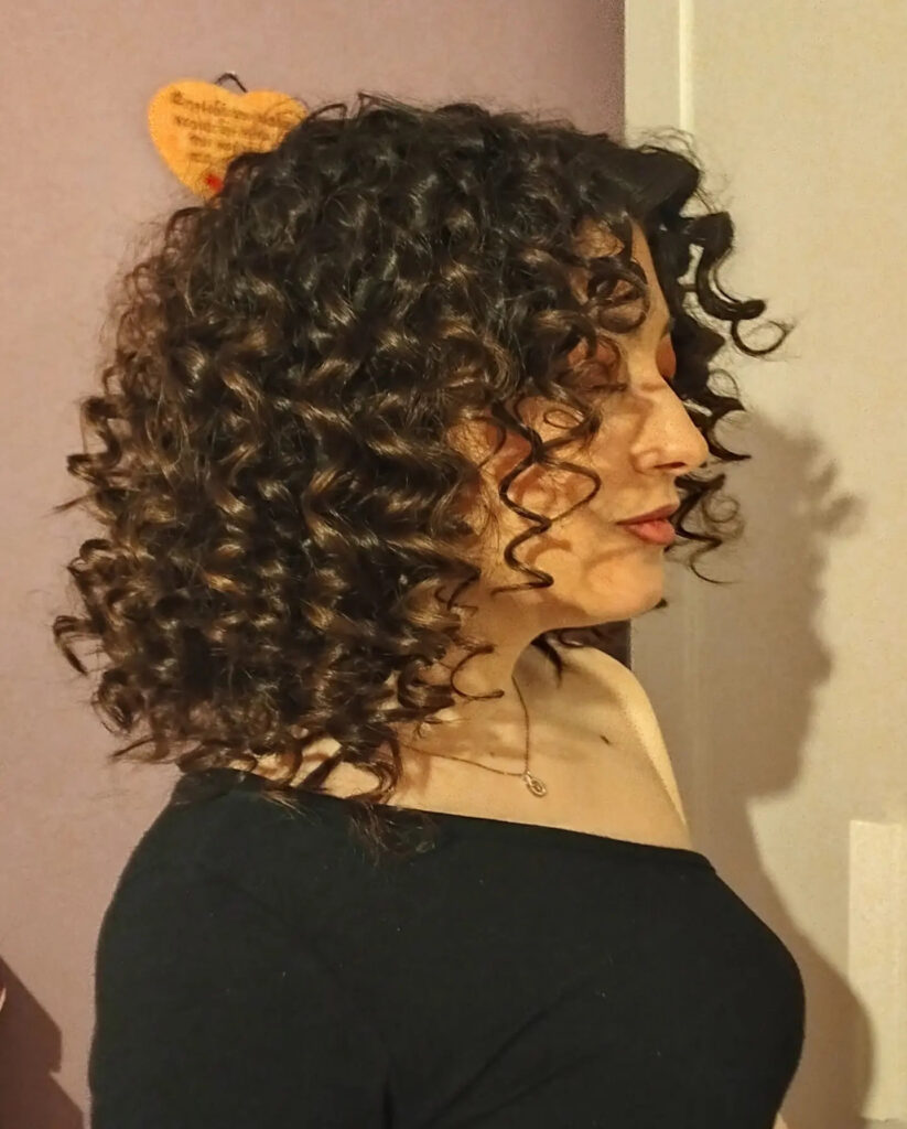 A side profile view of a woman with voluminous shoulder-length dark brown curls and subtle highlights.