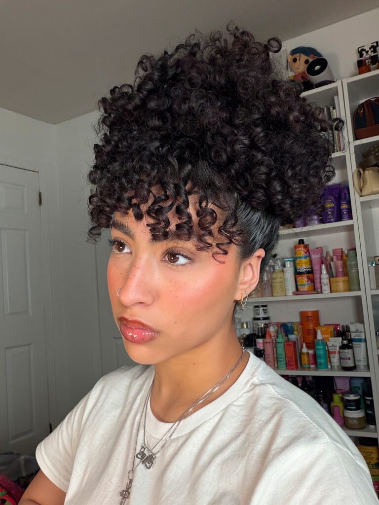 A woman with her dark curly hair styled in a high pineapple updo with defined curly bangs.