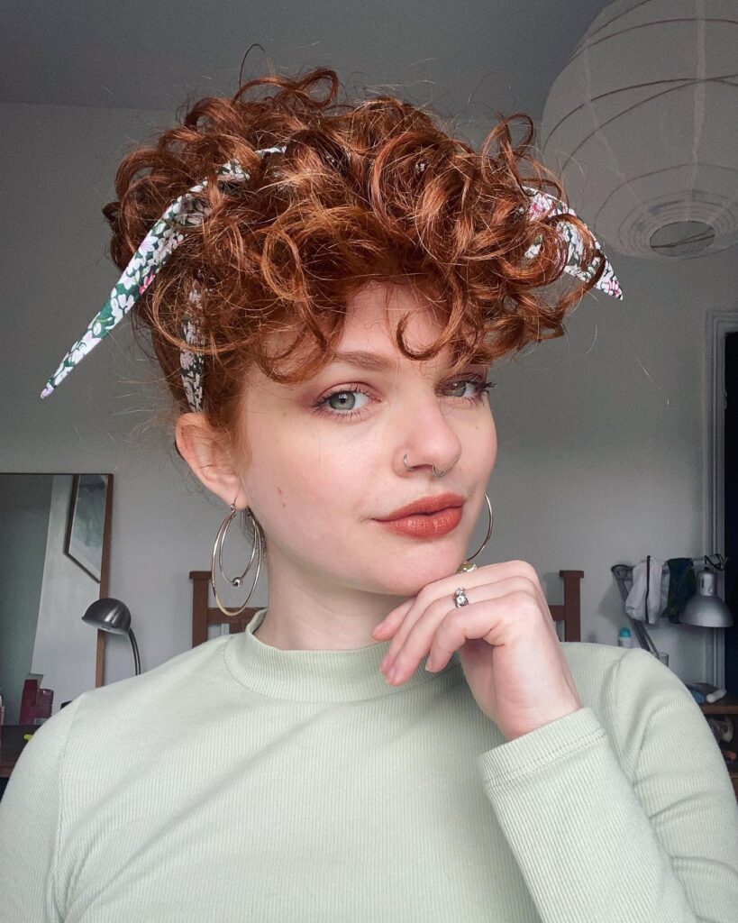 A woman with a short auburn curly pixie cut is wearing a floral-patterned headband.