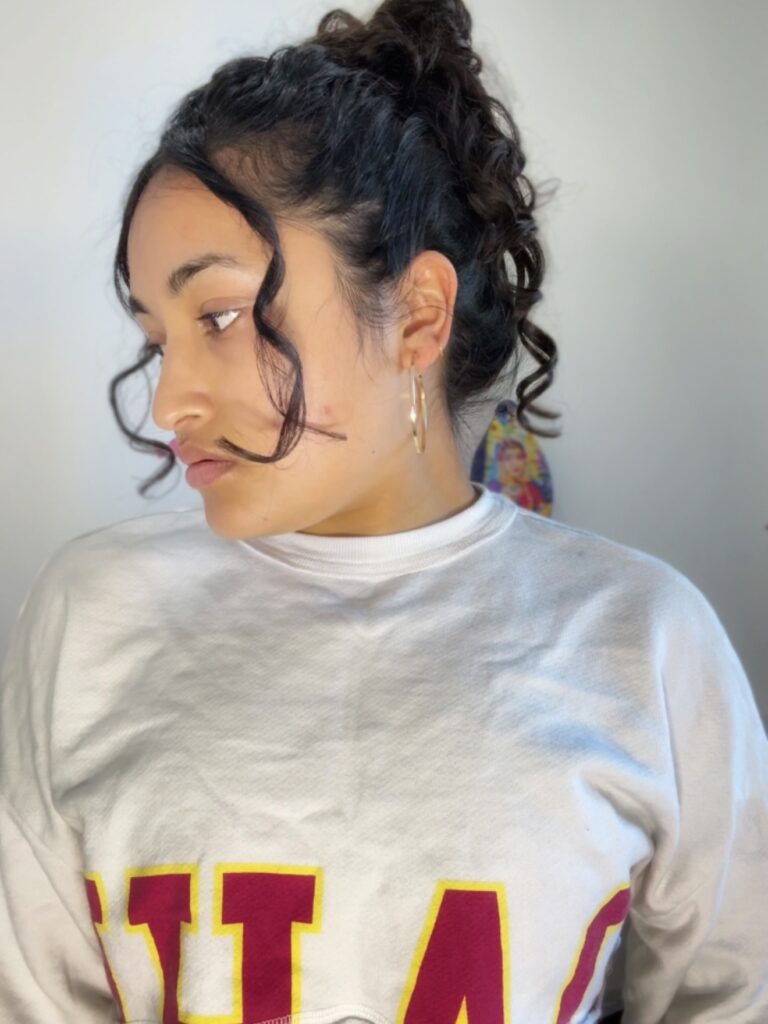 A woman with her dark curly hair styled in a high bun with long, thin curls framing her face.