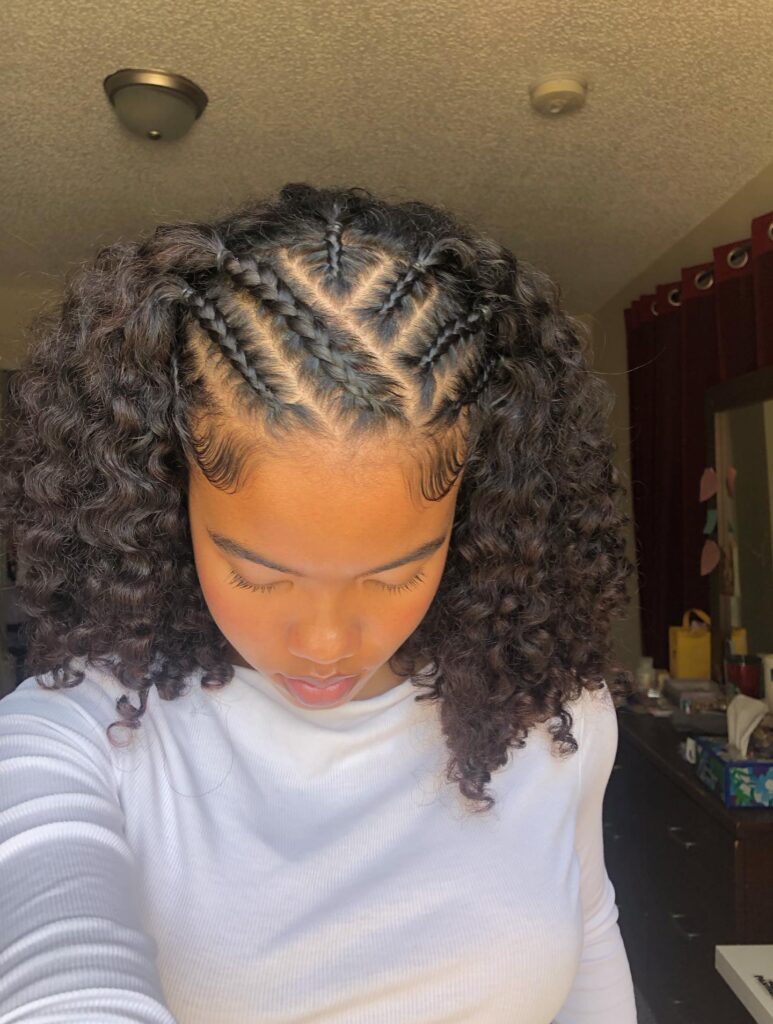 A woman with shoulder-length dark curls and several small diagonal braids styled across the top of her head.