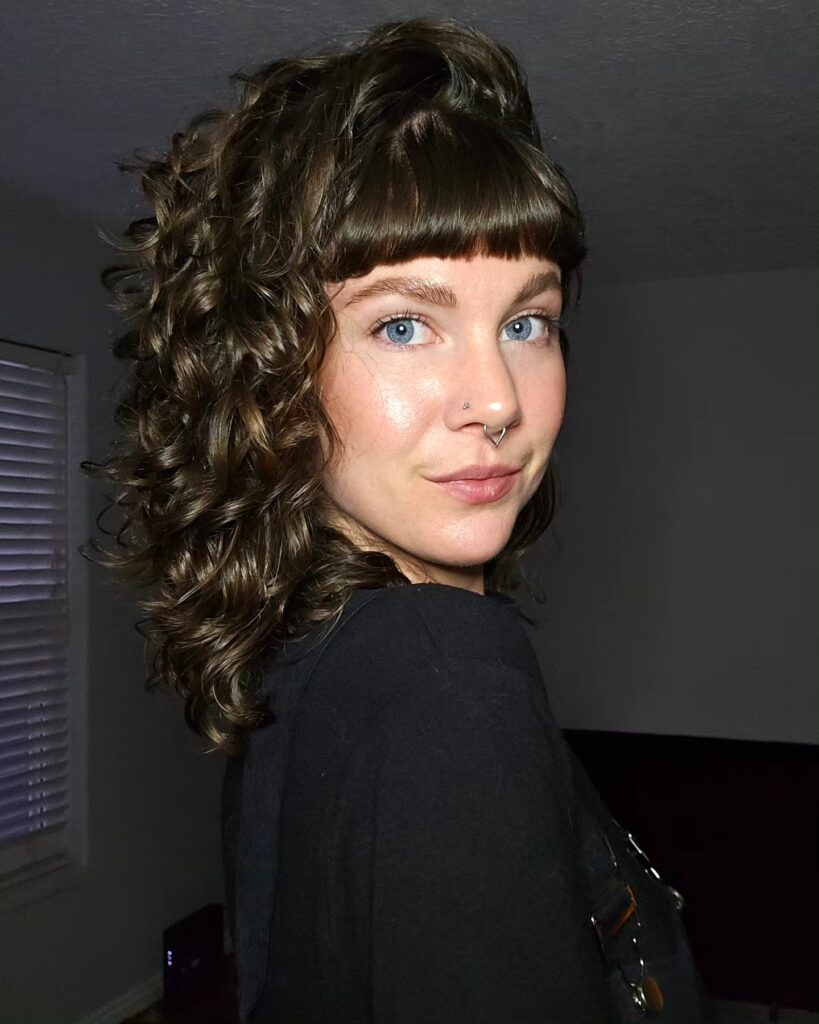 A woman with shoulder-length glossy brown curls and thick, blunt, straight bangs.