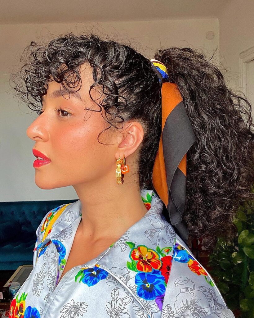 A side profile of a woman with a high curly ponytail tied with a patterned silk scarf and curly bangs.