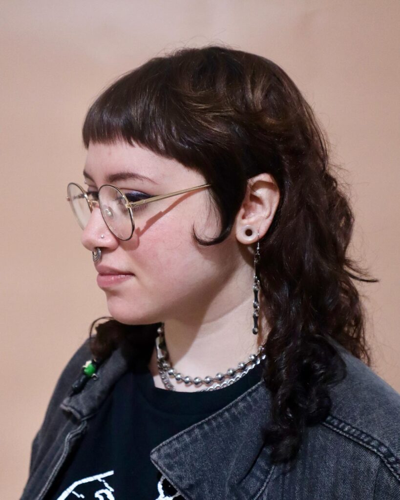 A woman with a medium-length curly mullet hairstyle featuring dark brunette hair and short, blunt baby bangs.