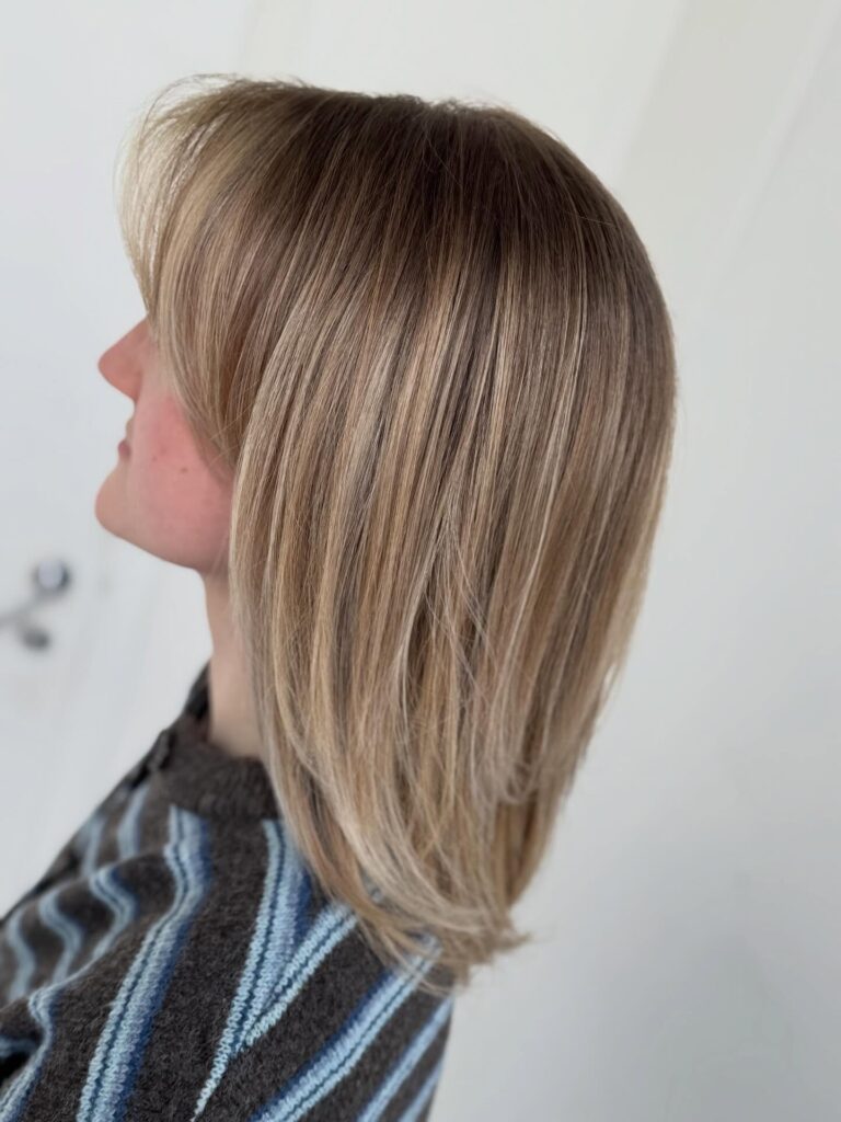 A side profile of a woman with a medium-length straight blonde lob featuring long wispy bangs and soft internal layers.